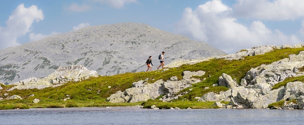 Two hikers walk on a trail by a mountain lake