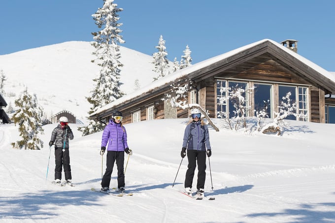 Family is skiing on a groomed slope outside a nice cabin, with mountains in the background