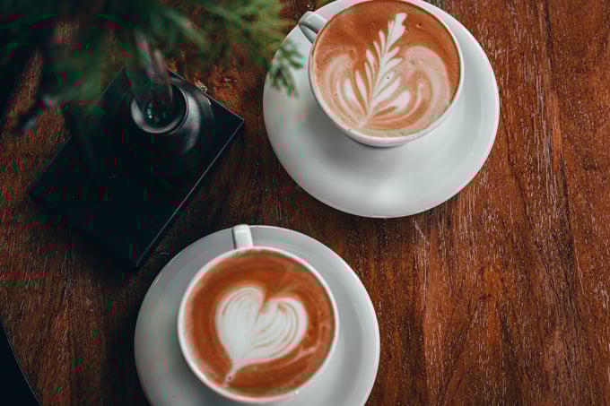 Two cups of cappuccino on a table, with heart and leaf patterns in the foam
